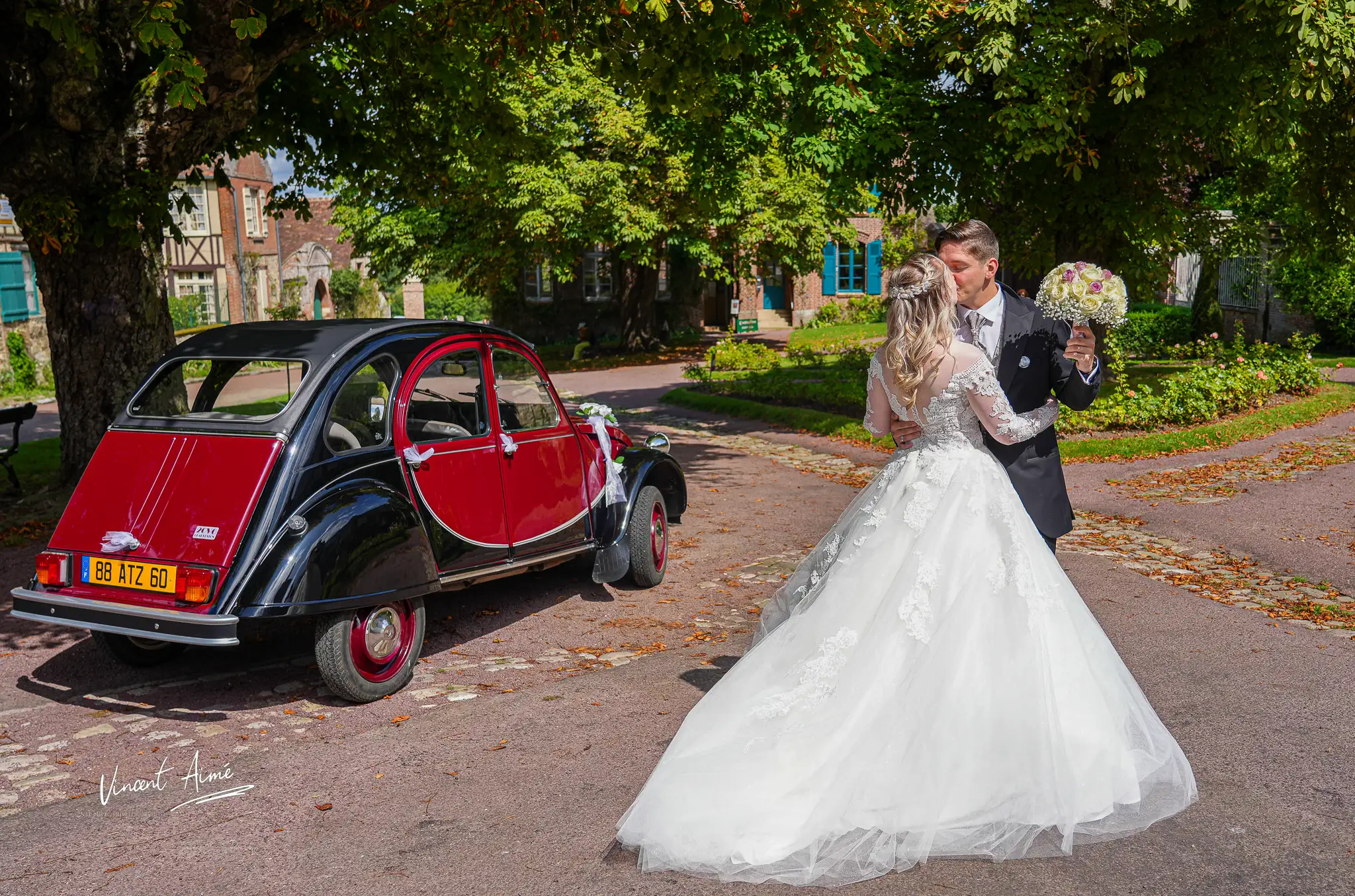 Deux mariés s'embrassent dans une allée devant une voiture ancienne