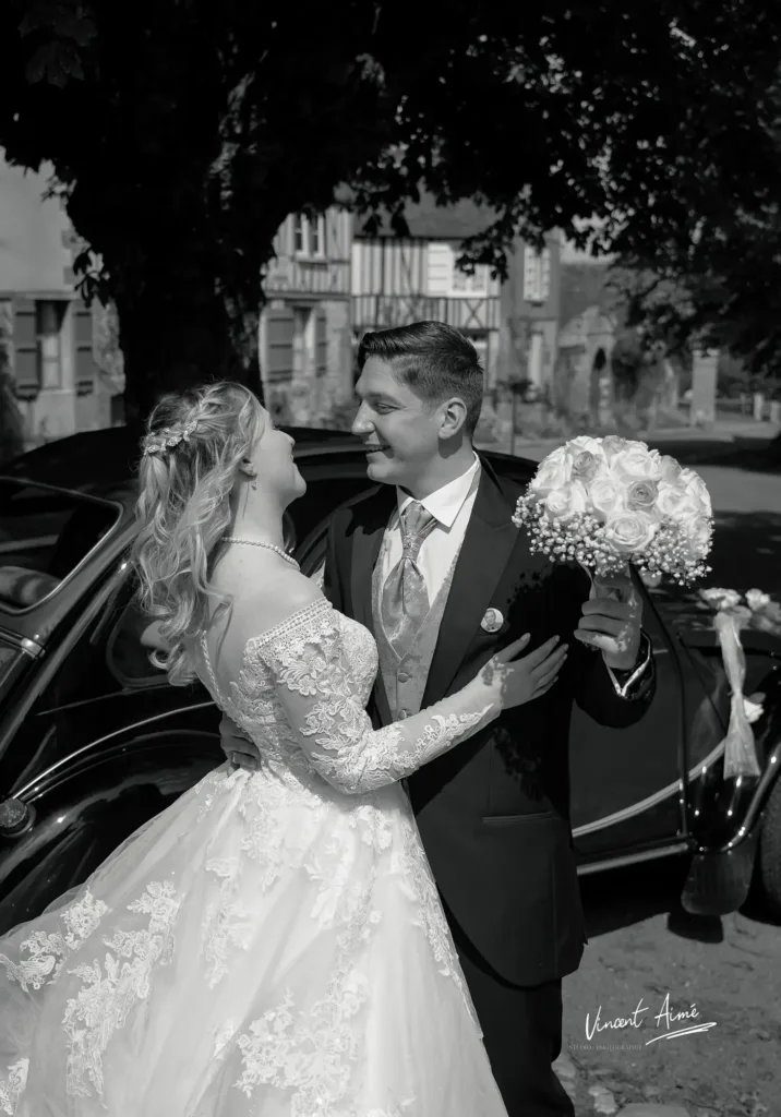 Marié tenant le bouquet et sa femme, devant leur voiture. Photo en noir et blanc