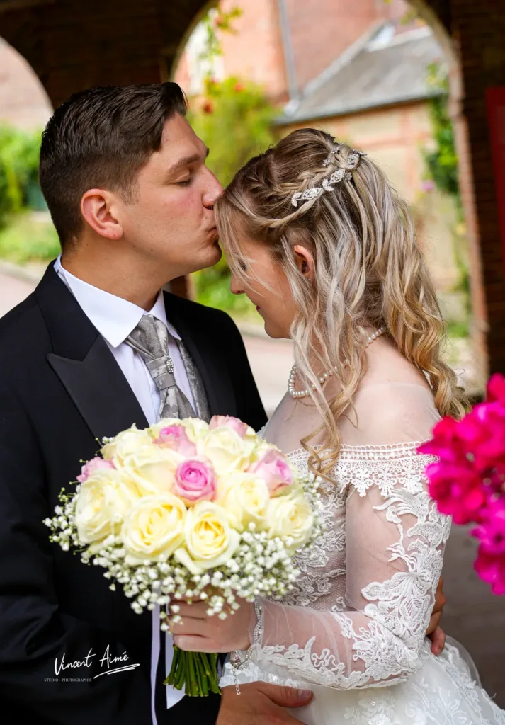 Le marié embrasse le front de la mariée. La mariée tient son bouquet de fleurs.