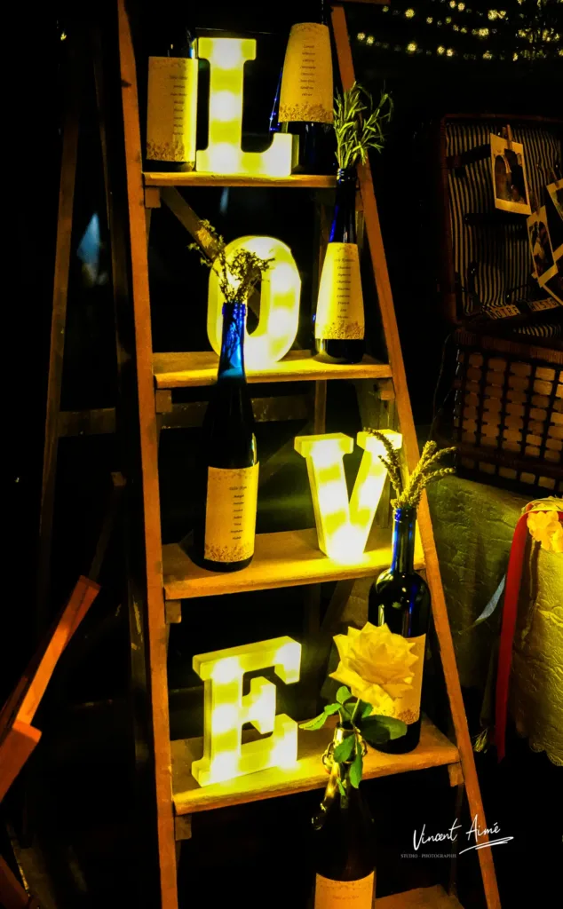 Etagère en bois avec des bouteilles contenant des fleurs, indiquant le plan de tables et des lettres éclairées formant le mot "LOVE"